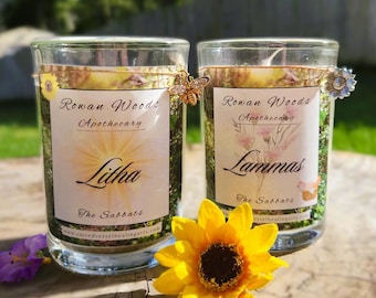 May include: Two glass candles with labels that read "Rowan Woods Apothecary." One candle is labeled "Litha" and the other "Lammas." The candles are decorated with a bee and a flower. A yellow sunflower and purple flower are in the foreground.