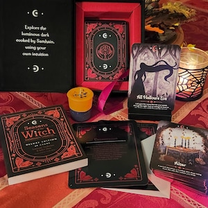 May include: A Seasons of the Witch Samhain Oracle deck in a black box with red accents. The set includes 60 cards with moth illustrations, a guidebook, and a card stand. One card displays a silhouette titled All Hallow's Eve, another shows an altar.