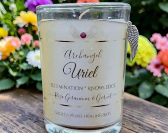 May include: A clear glass candle with a cream-colored wax. The candle has the words "Archangel Uriel" and "ILLUMINATION * KNOWLEDGE" printed on it. A silver-colored wing charm hangs from the rim. The candle is scented with Rose Geranium & Garnet.