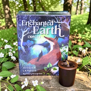 May include: A boxed deck of "Enchanted Earth Oracle" cards featuring a fox illustration. The box is dark blue with white text and a forest scene. A small brown candle with cloves sits in front of the box.