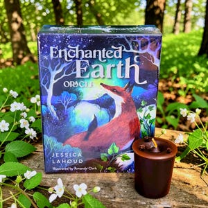 May include: An "Enchanted Earth Oracle" card deck box featuring a fox illustration, with the text "Enchanted Earth Oracle" and the names Jessica Lahoud and Amanda Clark. A small, dark brown candle sits next to the box.