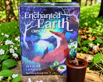 May include: An "Enchanted Earth Oracle" card deck box featuring a fox illustration, with the text "Enchanted Earth Oracle" and the names Jessica Lahoud and Amanda Clark. A small, dark brown candle sits next to the box.