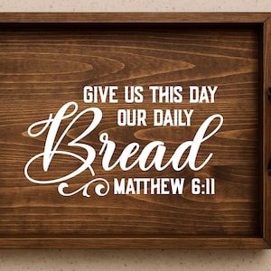 May include: A rectangular wooden serving tray with dark brown stain and black handles. The tray features white text that reads "GIVE US THIS DAY OUR DAILY Bread MATTHEW 6:11". The tray is designed for serving food or as a decorative piece.