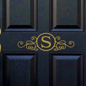 May include: A black front door with a gold door handle and a gold monogram decal with the letter "S" in a circle surrounded by decorative swirls.