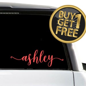 May include: A red vinyl decal with the name "ashley" in a cursive font. The decal is on the back window of a white car. A gold circle sticker with the text "BUY 1 GET 1 FREE" is on the car window.