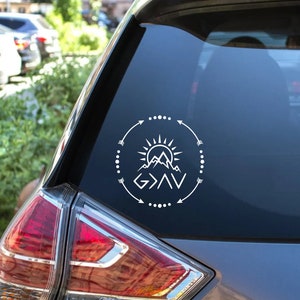May include: White vinyl decal with a mountain range, sun, and arrows forming a circle around the text "God is greater than the highs and lows".