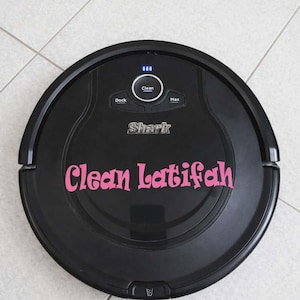 May include: A black Shark robot vacuum cleaner with the text "Clean Latifah" in pink script. The vacuum has buttons for "Dock", "Clean", and "Max". The vacuum is on a tiled floor.