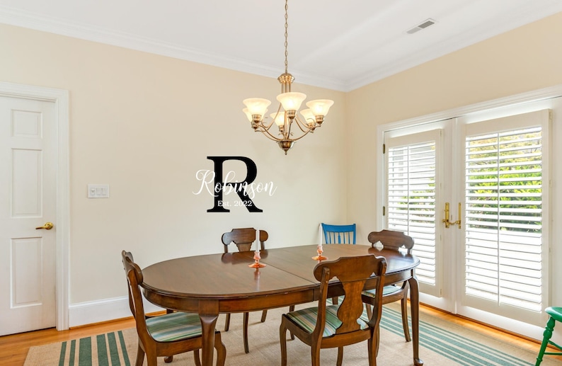 May include: A dining room with a wooden table and chairs. The table is oval shaped and has a dark wood finish. There are six chairs around the table, all with a similar dark wood finish. The room has white walls and a large window with white blinds. A black metal letter R with the text "Robinson Est. 2022" is on the wall.