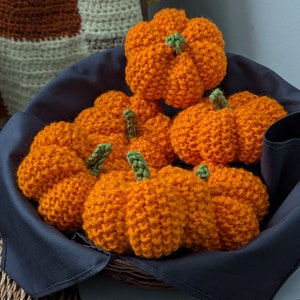 May include: A collection of handmade, orange crochet pumpkins with green stems, nestled in a dark blue basket. The pumpkins vary in size and are arranged to create a fall-themed display. The basket sits on a woven surface.