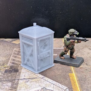 May include: A light gray miniature portable toilet and a miniature soldier figure with a rifle. The soldier is wearing a green and brown uniform and is standing on a black base. The toilet has a textured surface.
