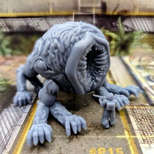 May include: A gray, detailed, 3D printed miniature of a monstrous creature with a large, gaping maw and multiple legs. The creature is posed as if attacking a smaller, gray, 3D printed miniature of a soldier holding a rifle.