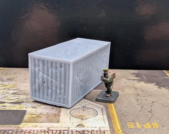 May include: A light gray miniature shipping container with vertical ridges. A small figurine of a person in a green uniform and hat stands nearby. The container is on a textured surface.