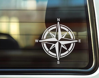 May include: White compass rose decal, featuring a star design with cardinal directions labeled N, E, S, and W. The compass is set within a circular frame. The decal is affixed to a car window.