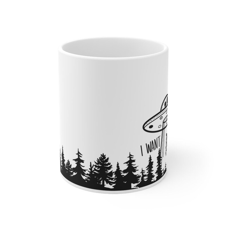May include: White ceramic mug with a black silhouette of a UFO flying over a forest with the text "I want to believe" printed on the mug.