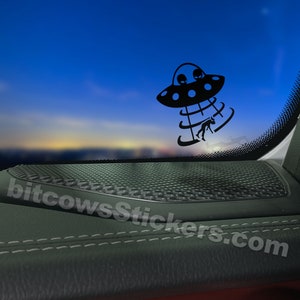 Puede incluir: Pegatina en blanco y negro de un OVNI secuestrando a una persona. La pegatina está colocada en un panel interior de coche negro con una rejilla de altavoz. El texto "bitcowsstickers.com" es visible en la esquina inferior derecha.