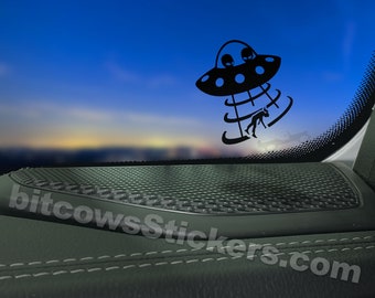 May include: Black and white sticker of a UFO abducting a person. The sticker is placed on a black car interior panel with a speaker grill. The text "bitcowsstickers.com" is visible in the bottom right corner.