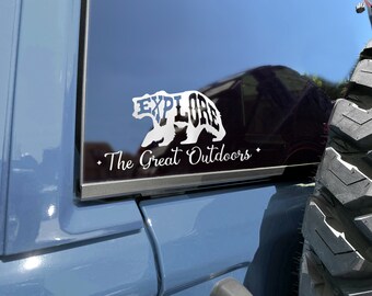 May include: A white vinyl decal on a blue car window. The decal is a silhouette of a bear with the word "EXPLORE" inside the bear's body. Below the bear is the text "The Great Outdoors".