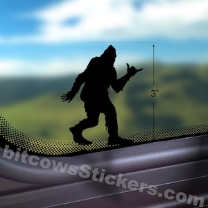 Op de afbeelding: Een zwarte silhouetsticker van een Bigfoot-wezen dat een shaka-handgebaar maakt. Een verticale meetlijn geeft aan dat de sticker 7,62 cm hoog is. De sticker is aangebracht op een glazen oppervlak met een wazig landschap op de achtergrond.