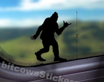 May include: A black silhouette sticker of a Bigfoot creature making a shaka hand gesture. A vertical measurement line indicates the sticker is 3 inches tall. The sticker is applied to a glass surface with a blurred landscape visible in the background.