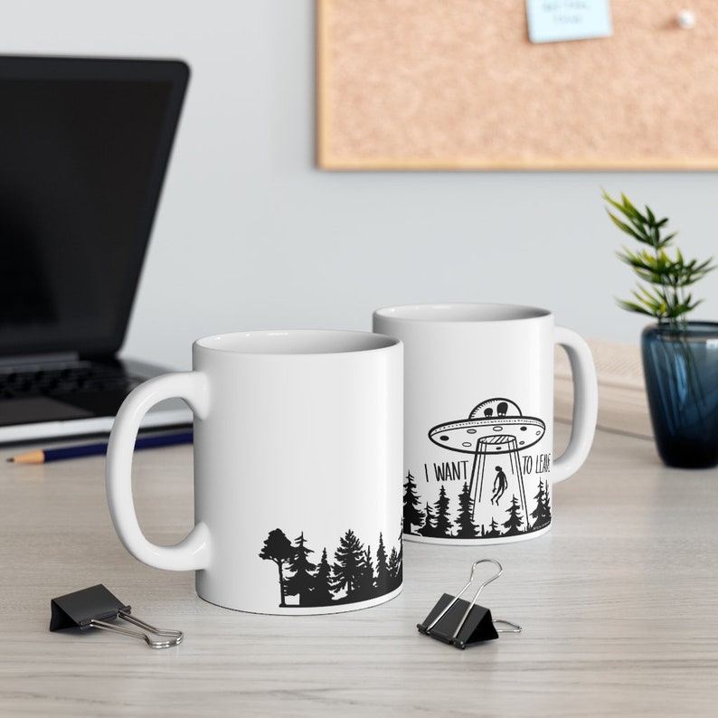 May include: Two white ceramic mugs with black silhouettes of a UFO, trees, and a person. The mug on the right has the text "I WANT TO LEAVE" printed on it.