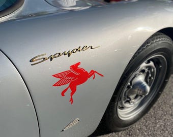 May include: A silver sports car fender featuring the gold script text Spyder and a red winged horse decal. The car has a black tire and a metallic wheel hub. A small silver emblem is positioned below the red winged horse graphic on the body.