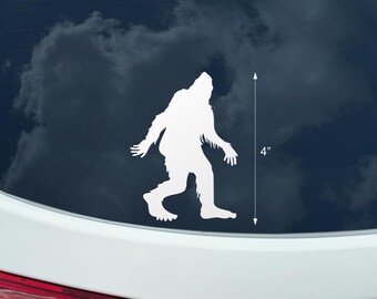 May include: A white vinyl decal of a Bigfoot silhouette, approximately 4 inches tall. The design depicts the legendary creature in a walking pose, with a shaggy appearance. The decal is shown against a dark background.