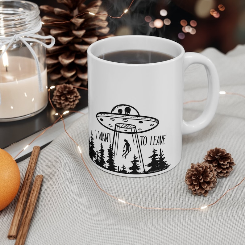 May include: White ceramic mug with a black and white illustration of a UFO, a person falling from the sky, and trees. The text "I want to leave" is printed on the mug.