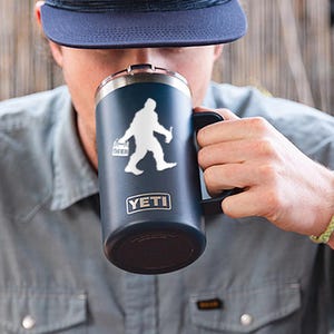 以下が含まれることがあります： 白いビッグフットがビール6本パックを持っている白いシルエットが描かれた、濃い青色のイエティランブラーマグ。マグには黒いハンドルが付いており、前面にはイエティのロゴが印刷されています。