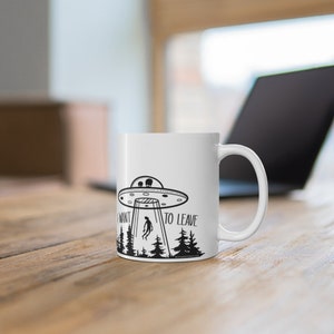 May include: White ceramic mug with a black and white illustration of a UFO abducting a person in a forest. The text on the mug reads "I want to leave".