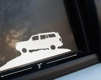 May include: A white SUV sticker, approximately 3 inches in length, is depicted on a window. The sticker shows a side profile of the vehicle climbing a hill. The background is a blurred view of the outdoors.