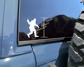 May include: White vinyl sticker of a Bigfoot silhouette giving a thumbs up. The sticker is on a blue car window and measures 5 inches tall.