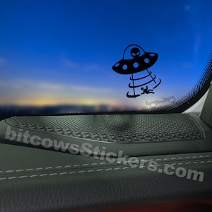 May include: Black vinyl sticker featuring a cartoon UFO with an alien and a person being abducted. The UFO is black with blue accents. The sticker is applied to a car window, with the website bitcowsStickers.com visible.