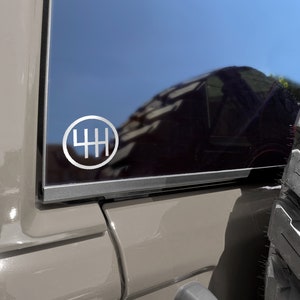 Puede incluir: Primer plano de un vehículo gris con una pegatina 4H plateada en la ventana trasera. La pegatina es un círculo con un patrón de cambio de marchas en su interior.