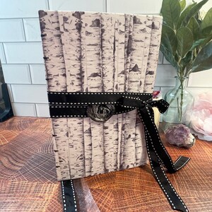 May include: A white journal with a birch tree print cover, tied with a black ribbon with white stitching. The ribbon has a decorative metal seal.
