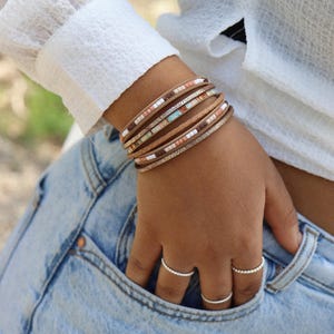 May include: A stack of leather bracelets with colourful beads and metallic accents. The bracelets are worn on a person's wrist.