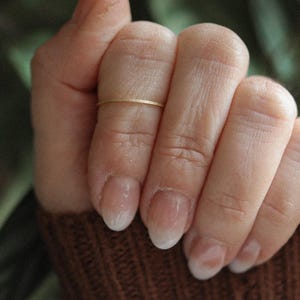 May include: A close-up of a hand wearing a thin gold band ring. The hand is resting on a brown knitted fabric.