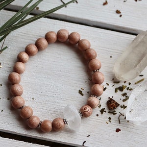 May include: A wooden beaded bracelet with a clear quartz crystal charm. The bracelet is on a white wooden surface with dried herbs and two clear quartz crystals.