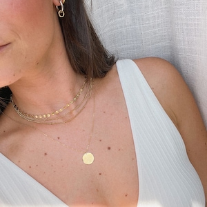 May include: A woman wearing a white vest top with a gold layered necklace featuring a round pendant.