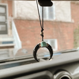 May include: A black crescent-shaped car charm with a turquoise stone accent, suspended by a black cord. The charm is hanging from a car's rearview mirror, with a blurred background of a house and street.