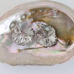May include: A pair of silver-toned hoop earrings featuring a textured, fan-like pleated design. The earrings have a polished metallic finish and include clear plastic earring backs for secure fastening, showcasing a distinct radial pattern.