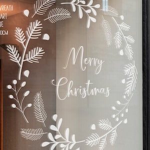 Puede incluir: Pegatina de vinilo blanco con un diseño de corona de Navidad y el texto "Feliz Navidad" en una ventana de vidrio.