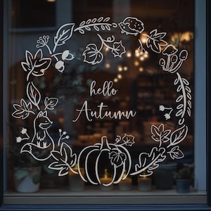 Pode incluir: Autocolante de janela branco com tema de outono, apresentando um design de coroa circular. A coroa inclui folhas, uma abóbora, uma raposa e o texto "hello Autumn". O autocolante é exibido numa janela com um fundo desfocado.
