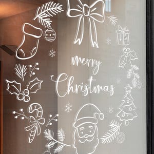 Könnte beinhalten: Weißes Weihnachtsfenster-Dekor mit einem Kranzdesign mit Schleife, Strumpf, Geschenk, Weihnachtsmann, Zuckerstange und Weihnachtsbaum. Die Worte "merry Christmas" stehen in der Mitte des Kranzes.