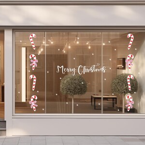 K&ouml;nnte beinhalten: Ein Schaufenster, das zu Weihnachten mit Zuckerstangen- und Schleifenaufklebern dekoriert ist. Die Worte "Merry Christmas" sind in Schreibschrift geschrieben. Wei&szlig;e Schneeflockenaufkleber sind ebenfalls vorhanden.
