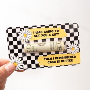 Puede incluir: Una tarjeta a cuadros en blanco y negro con un dise&ntilde;o de margarita amarilla. La tarjeta tiene una ventana de pl&aacute;stico transparente que muestra un billete de d&oacute;lar enrollado. El texto en la tarjeta dice "Iba a darte un regalo, pero luego record&eacute; que el efectivo es mejor."