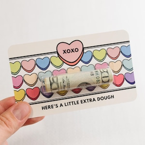 Puede incluir: Una tarjeta blanca con un coraz&oacute;n rosa que dice "XOXO" y una fila de corazones de caramelo coloridos. La tarjeta dice "Here's a little extra dough" y tiene un billete de d&oacute;lar enrollado dentro.