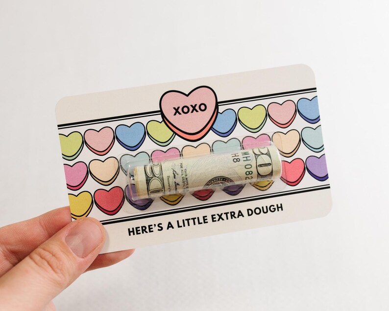 Puede incluir: Una tarjeta blanca con un coraz&oacute;n rosa que dice "XOXO" y una fila de corazones de caramelo coloridos. La tarjeta dice "Here's a little extra dough" y tiene un billete de d&oacute;lar enrollado dentro.