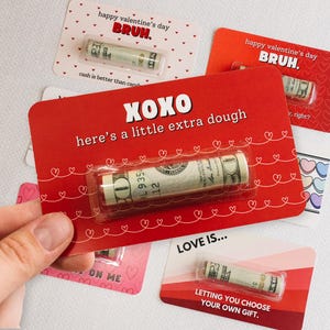 Puede incluir: Cuatro tarjetas del D&iacute;a de San Valent&iacute;n con corazones rojos y rosas y el texto "happy valentine's day BRUH." y "XOXO here's a little extra dough" y "LOVE IS... LETTING YOU CHOOSE YOUR OWN GIFT." Cada tarjeta tiene un billete de d&oacute;lar enrollado en su interior.
