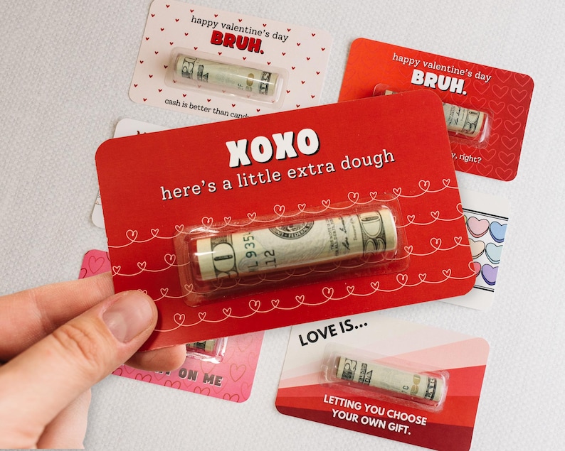 Puede incluir: Cuatro tarjetas del D&iacute;a de San Valent&iacute;n con corazones rojos y rosas y el texto "happy valentine's day BRUH." y "XOXO here's a little extra dough" y "LOVE IS... LETTING YOU CHOOSE YOUR OWN GIFT." Cada tarjeta tiene un billete de d&oacute;lar enrollado en su interior.