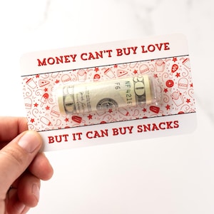 Puede incluir: Una tarjeta blanca con texto rojo que dice "El dinero no puede comprar el amor, pero puede comprar bocadillos." Se puede ver un billete de d&oacute;lar enrollado en la tarjeta.